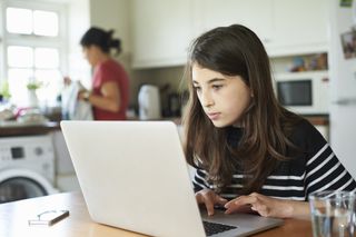Tween girl on a laptop at home in the kitchen while her mother is at the sink in the background