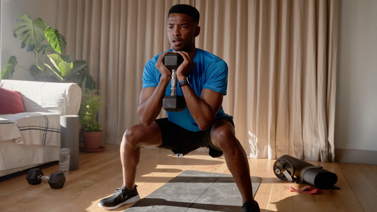 Young African American male exercising from home concentrating while squatting with a dumbbell in living room, workout at home