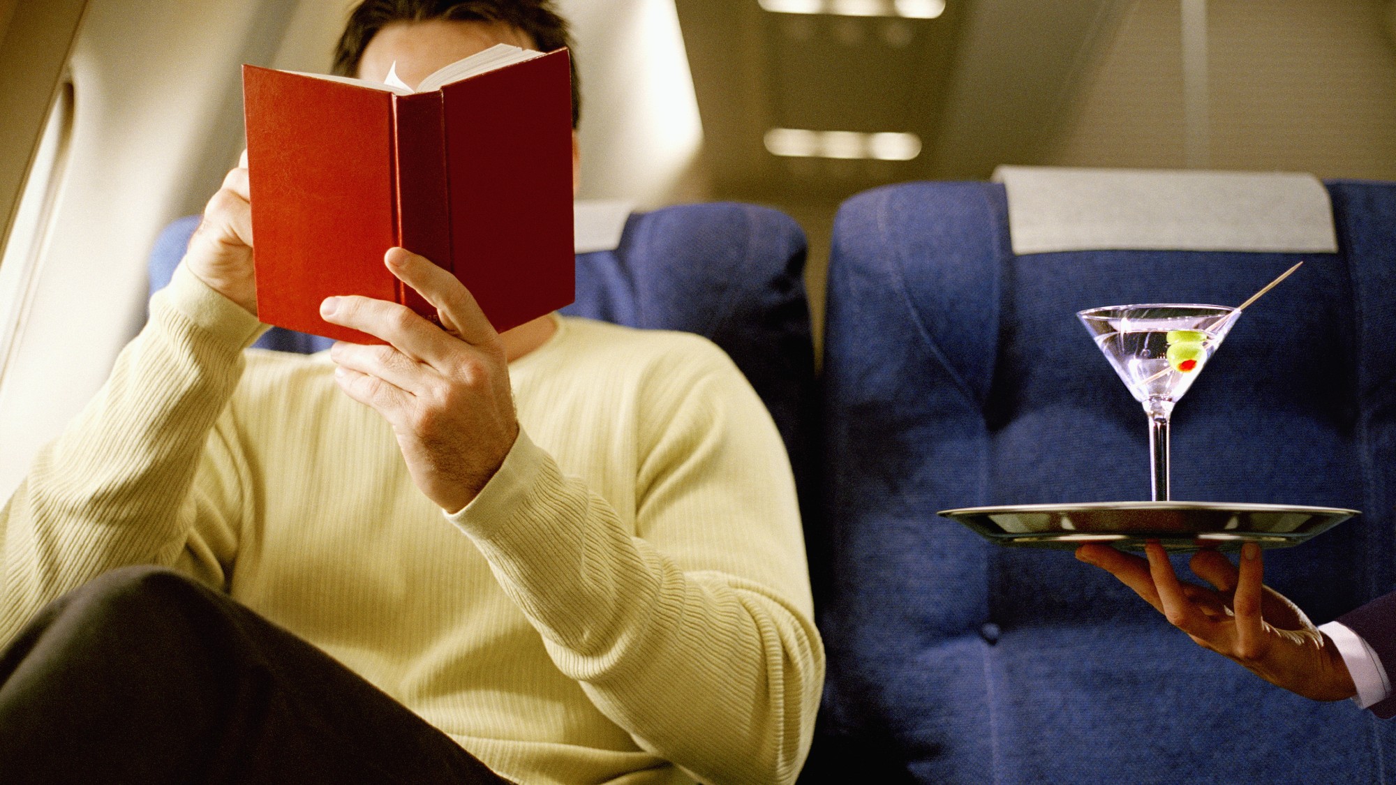 Man sitting in first class on an airplane reading a book and being served a martini