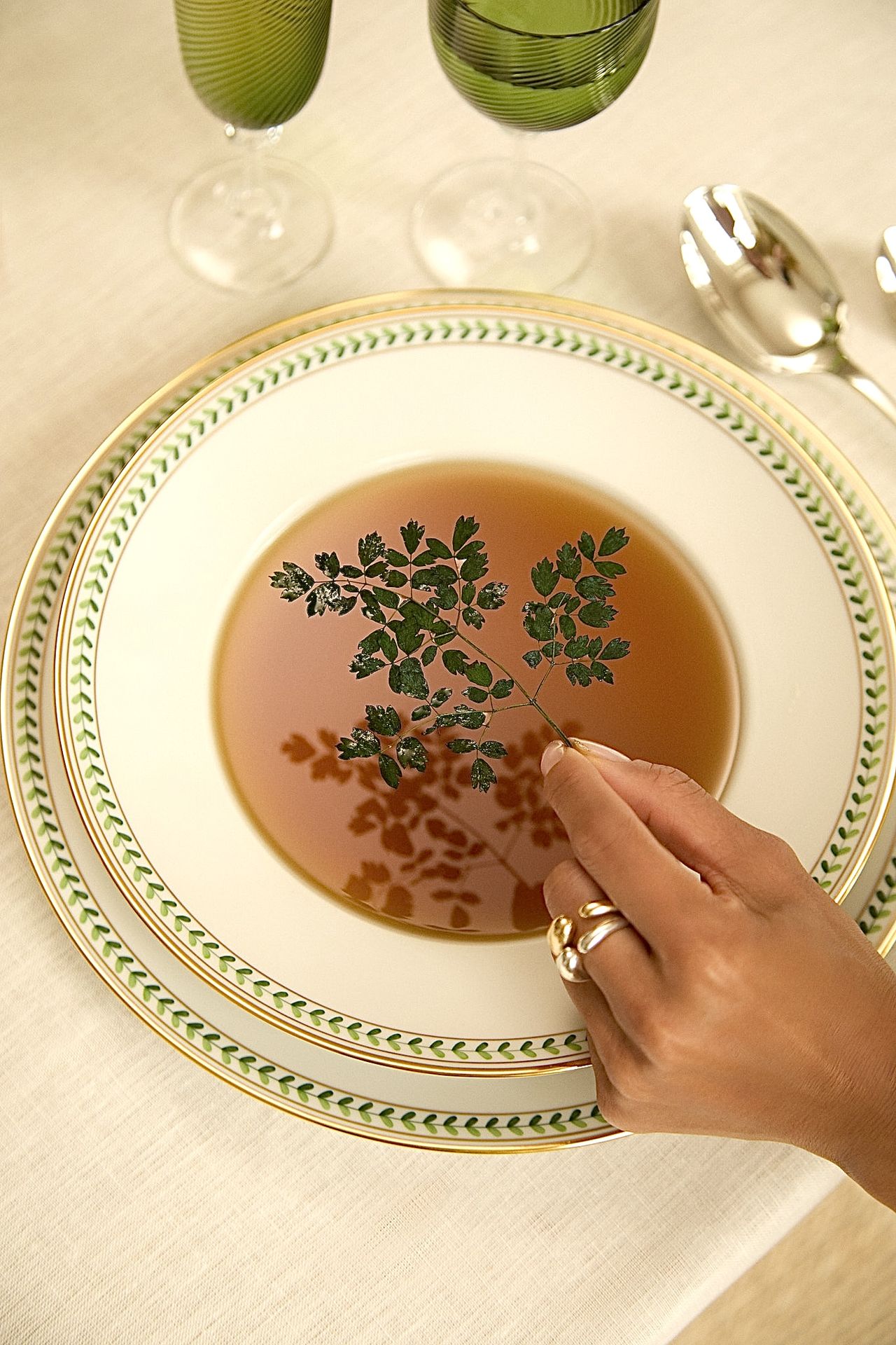 The hand of a woman, decorated with two rings, hangs over a dish filled with a red-hued soup as she holds a leafy string over it that casts a shadow on its contents.