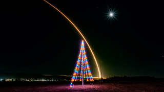 A SpaceX Falcon 9 rocket streaks over a tree-like multi-colored light decoration with the moon overhead.