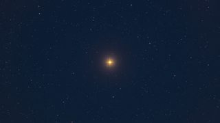 A bright yellow star, shining rays spiked on its sides to from a small "+", hangs in the center of a rich, dark blue, almost black sky. Fainter stars are sprinkled throughout.