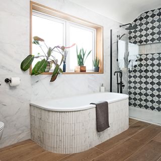 a curved bathtub under a window in a bathroom, clad in pencil-effect marble tiles and a row of houseplants on the windowsill above