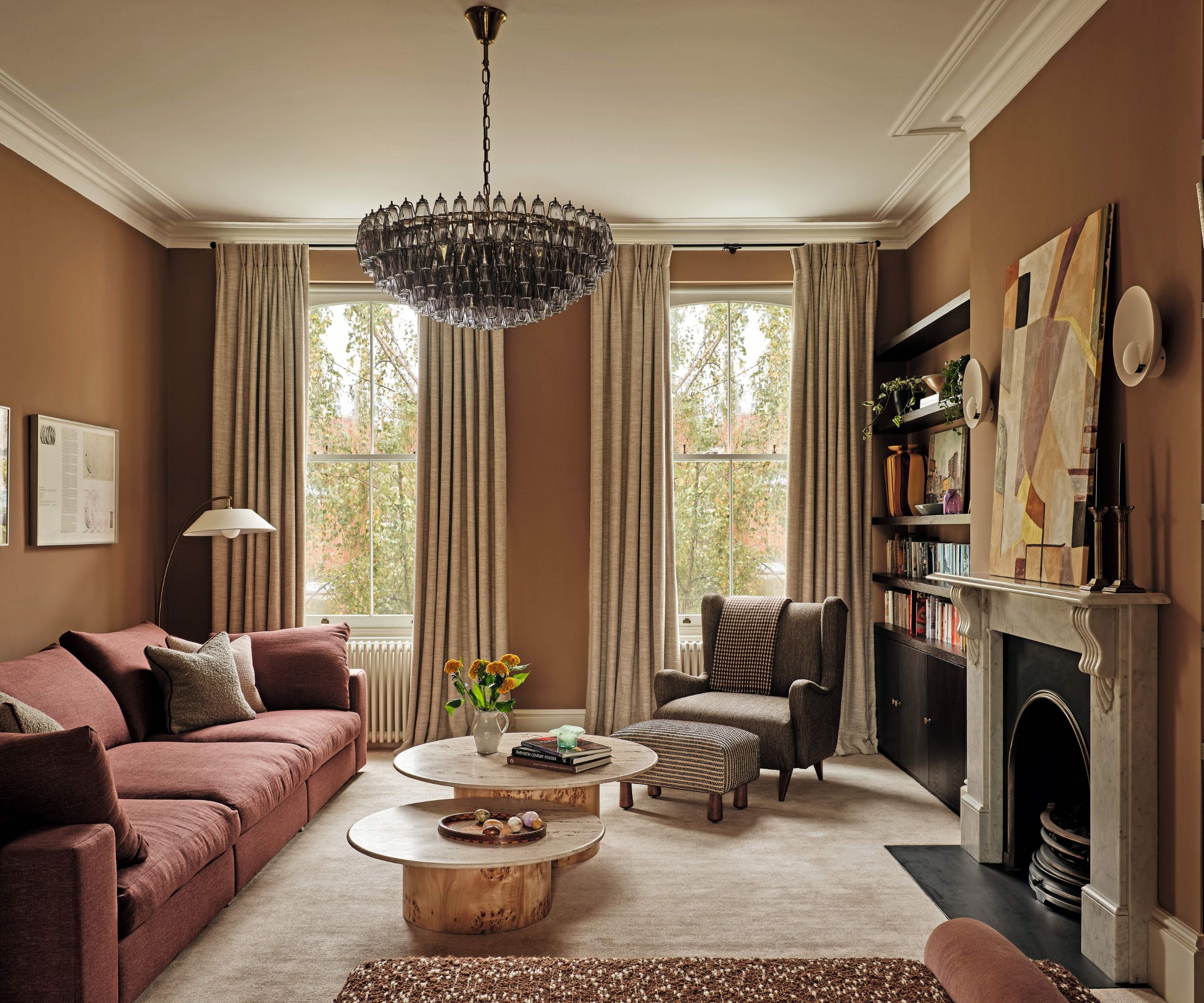 A warm, earth-toned living room featuring terracotta walls, a dusty rose sectional sofa, and a large tiered glass chandelier. Two organic-shaped wooden coffee tables sit on a plush beige rug before a marble fireplace.