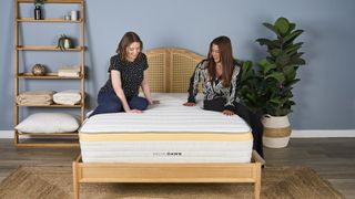Two women sit on the Helix Dawn Mattress for stomach sleepers, photographed on a light wooden bed frame in a blue bedroom