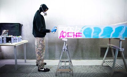 Spray booth, metal grids on a concrete floor, spray mask, man in black hooded sweater, combat pants, black trainers, blue plastic gloves using spray paint, aart work on wooden stands, side wodd and metal leg table with black suitcase and cans of spray paint, white back wall