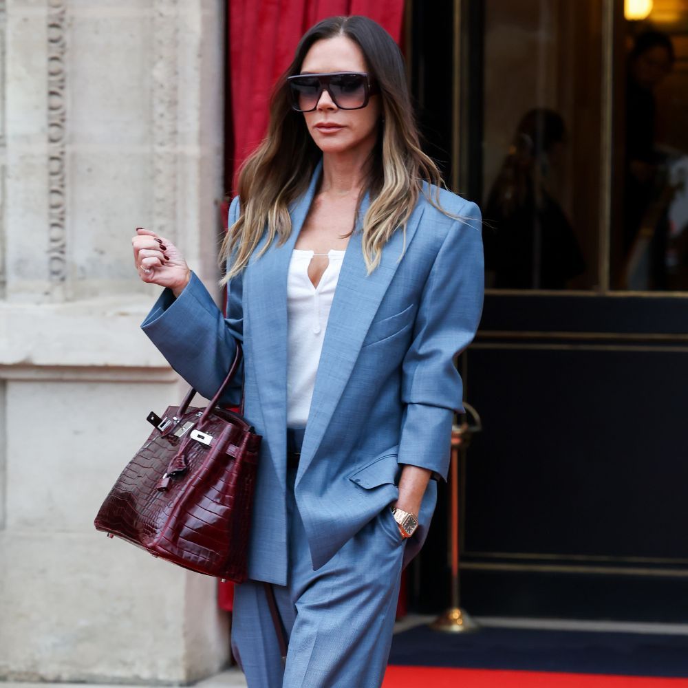Victoria Beckham wears a light blue suit with a dark burgundy bag and large aviator sunglasses.