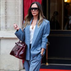 Victoria Beckham wears a light blue suit with a dark burgundy bag and large aviator sunglasses.