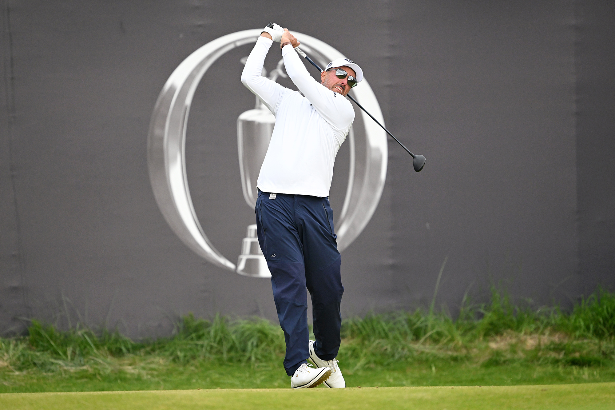 Phil Mickelson in the finish position after hitting a driver off the first tee during the opening round of the Open Championship at Royal Portrush, in front of the grandstands with the Open logo