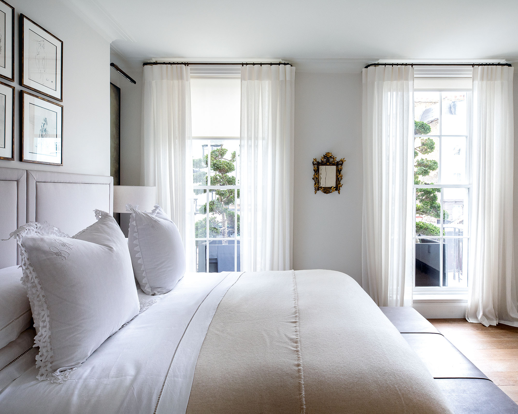 A bright, airy bedroom featuring a bed with crisp white linens, lace-trimmed pillows, and a cream-colored throw. The room has light grey walls, two large windows with sheer white curtains, and a small ornate gold mirror hanging between them. Four framed sketches are displayed on the wall above the upholstered headboard.