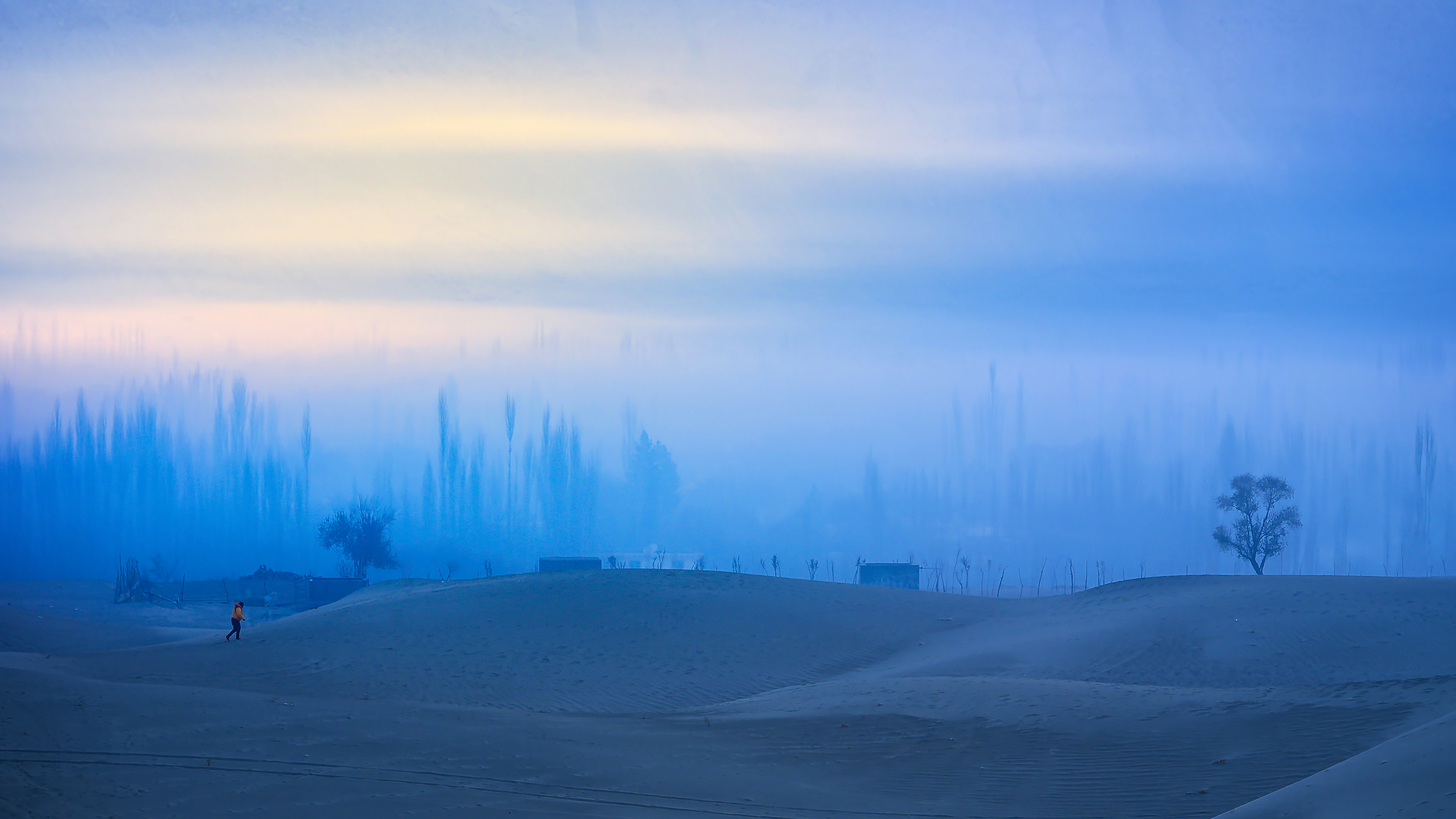 A lone figure walks on soft sand dunes under a blue, misty sky. Sparse trees and distant structures create a serene, dreamlike landscape at dawn