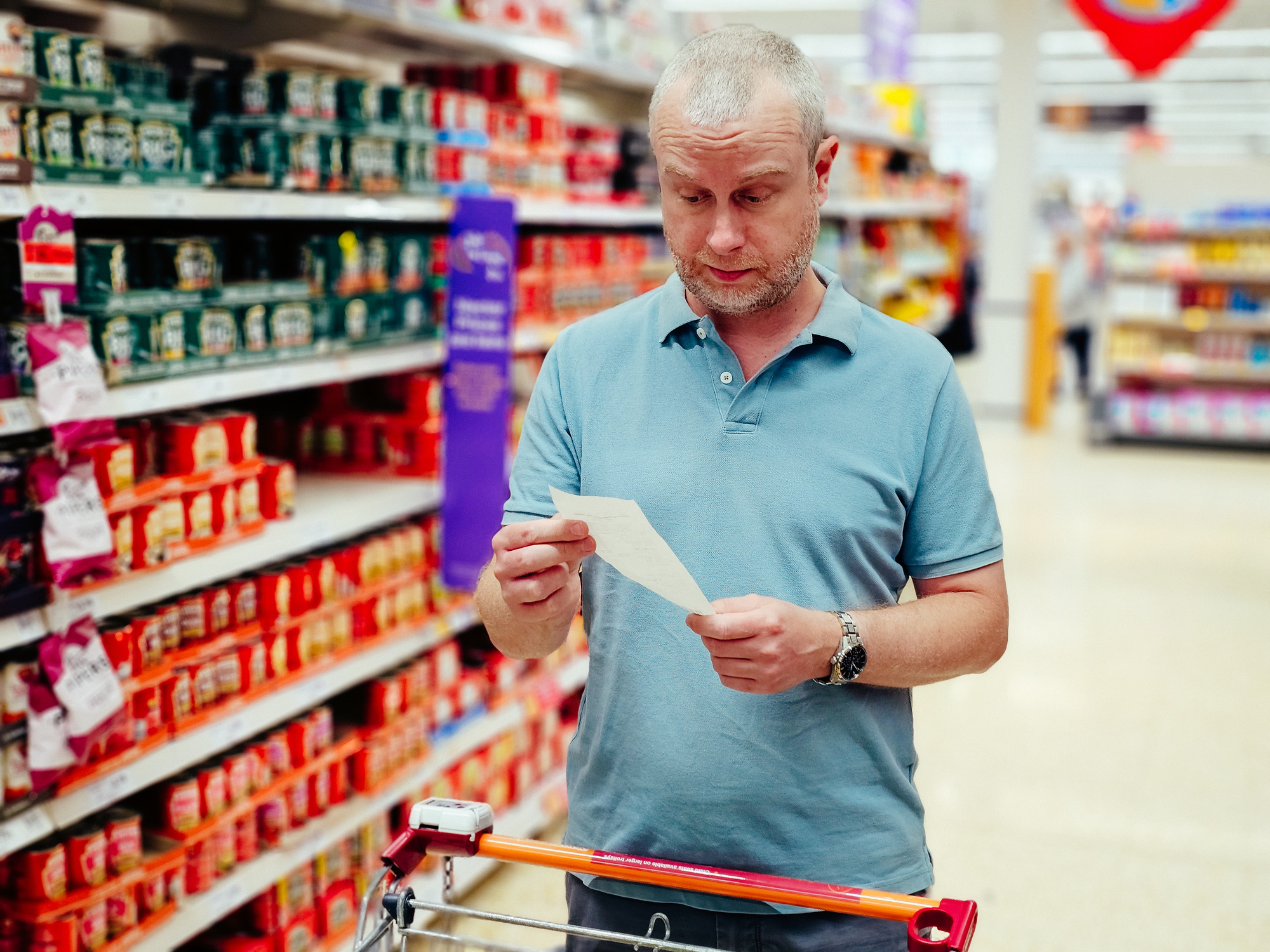 Color image depicting a mid adult man checking his shopping list in the supermarket aisle with a concerned face based on rising prices and inflation.