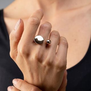 A close-up photograph of a hand shows a woman wearing a golden, abstract and organic-shaped ring with two spherical ends as she holds her wrist in front of her chest, which is dressed in a black shirt.