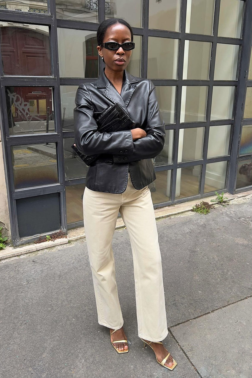 A woman wearing a black leather blazer, white jeans, white sandals, and black sunglasses.