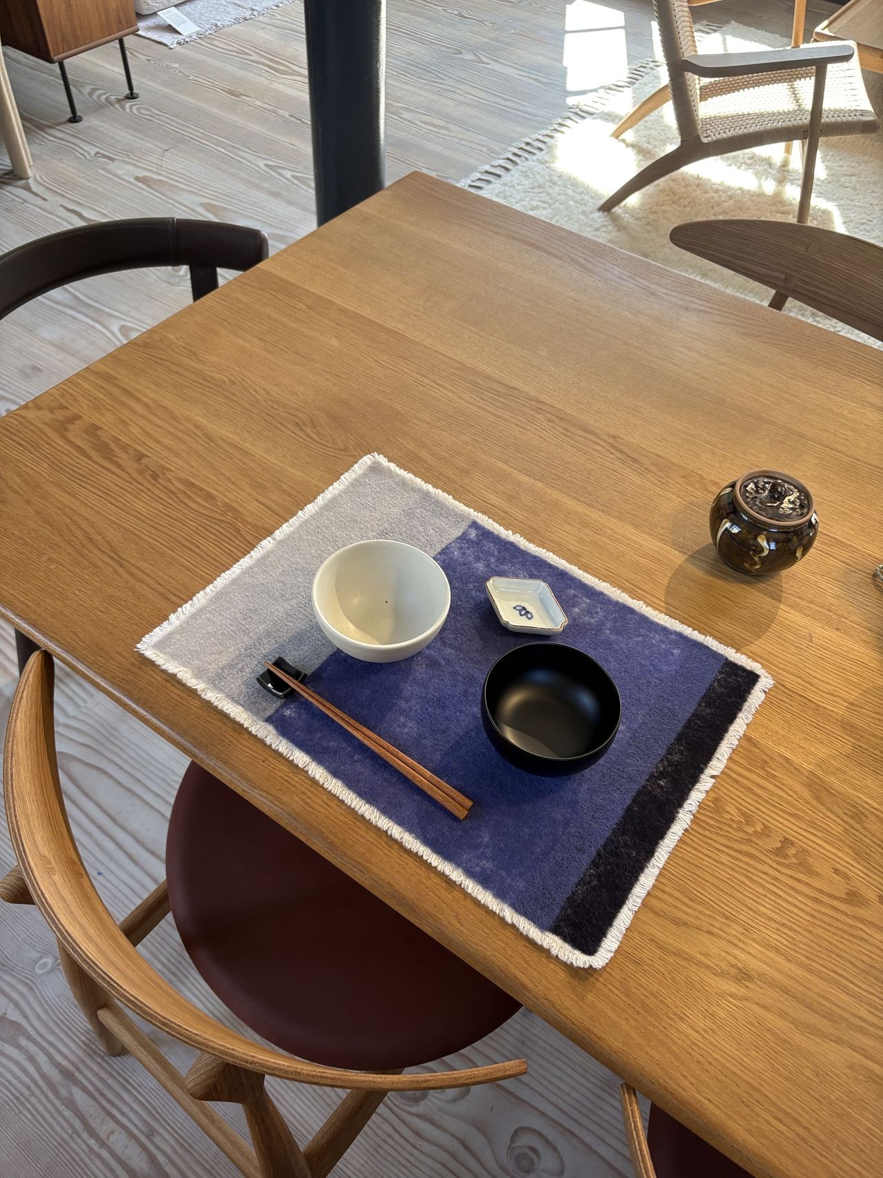 Image of a wooden table with a Japanese placemat at one of the seats with a black and a white bowl on it and chopsticks.