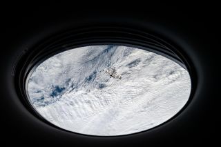 Through a horizontal ovular window, a section of Earth can be seen below, mostly covered in white clouds. Below, above the sky, a small cross frame can be seen - the international space station.