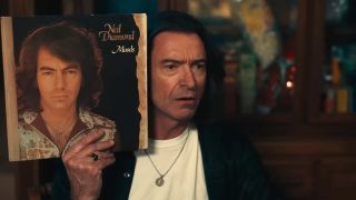 Hugh Jackman as Mike Sardina holds a Neil Diamond up to his head for comparison in the trailer for Song Sung Blue.