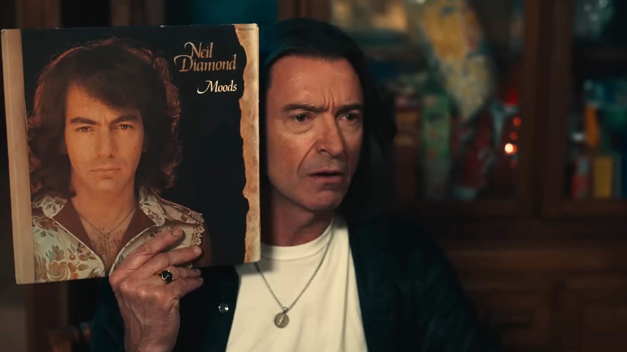 Hugh Jackman as Mike Sardina holds a Neil Diamond up to his head for comparison in the trailer for Song Sung Blue.