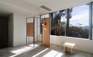 View of a black framed room divider with two wood panels and lighting attached. There is also a small light coloured trolley in a space with white walls, grey floors and windows offering a view of the trees and sky outside
