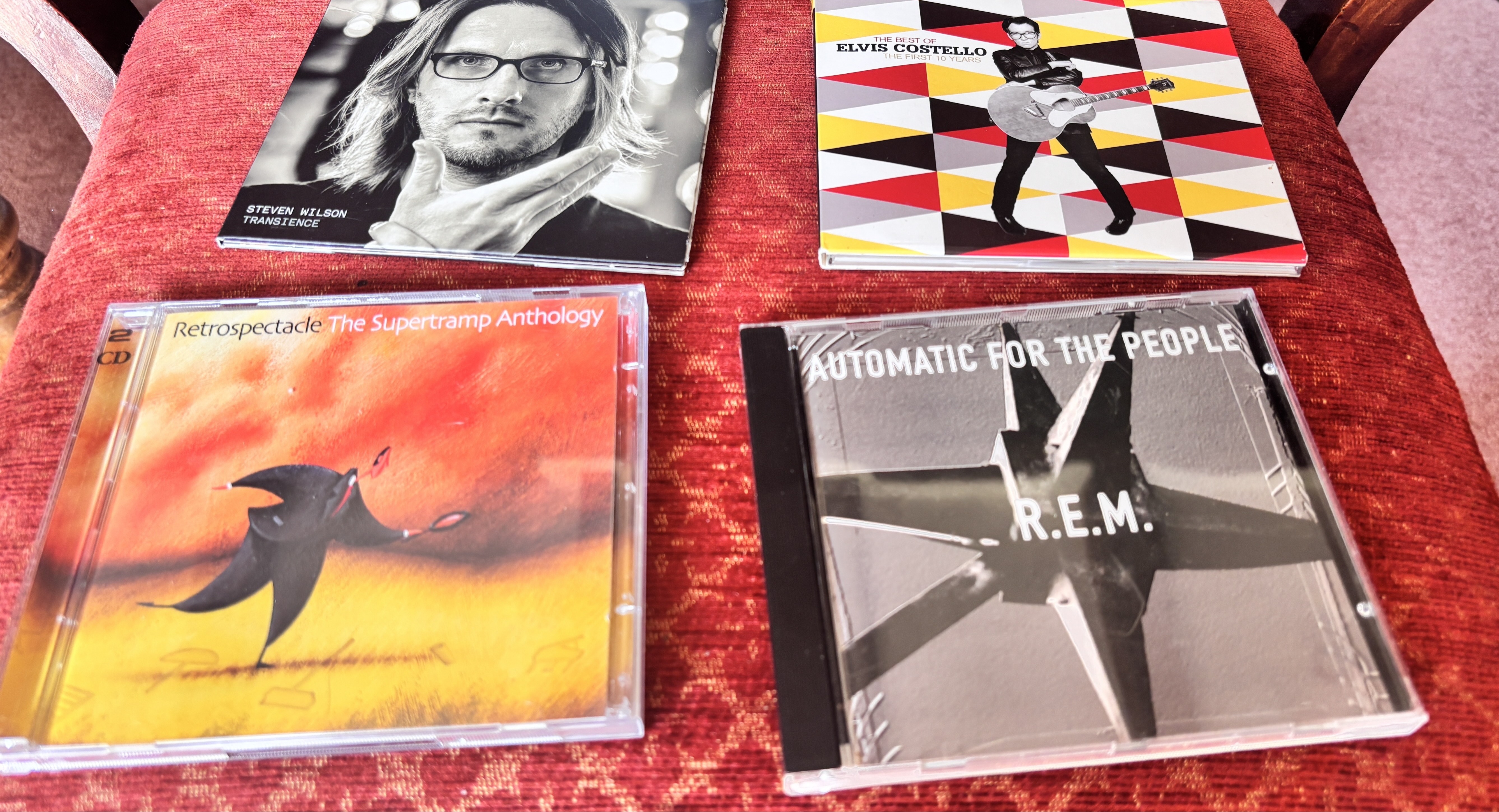 Four CDs on a red fabric surface including REM and Elvis Costello