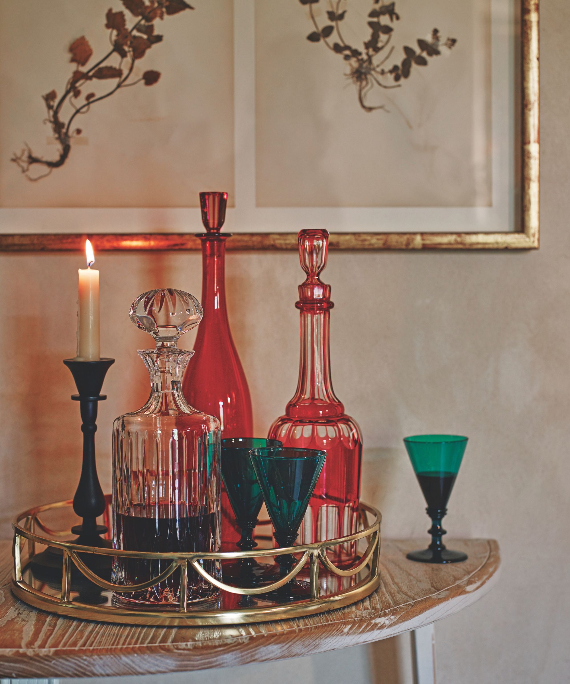 Vintage colored glassware on a gold mirrored tray on a wooden table. There is a lit taper candle, and above are two vintage-look botanical prints in a gold frame.