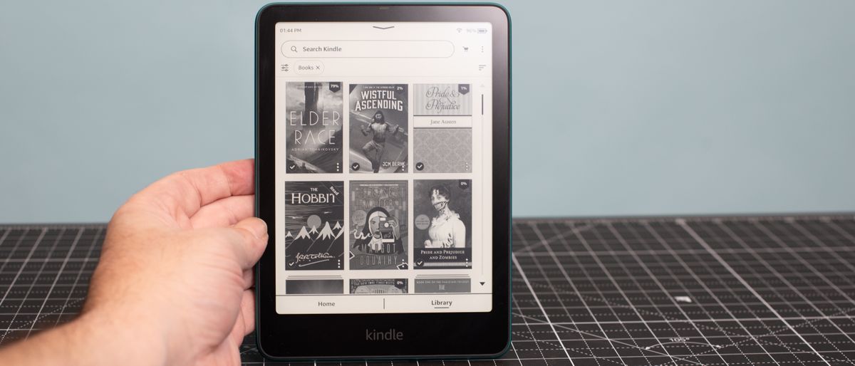 Kindle Paperwhite held up by one hand showing book covers in a grid including Elder Race by Adrian Tchaikovsky and Wistful Ascending by JCM Berne and The Hobbit by J.R.R. Tolkien