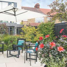A small garden patio with tables and chairs, a sun umbrella, dense planted flower borders and flowers.