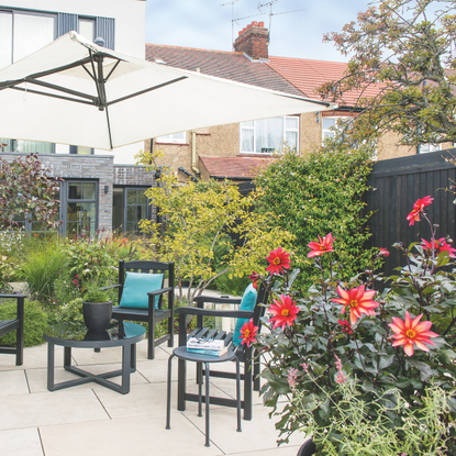 A small garden patio with tables and chairs, a sun umbrella, dense planted flower borders and flowers.