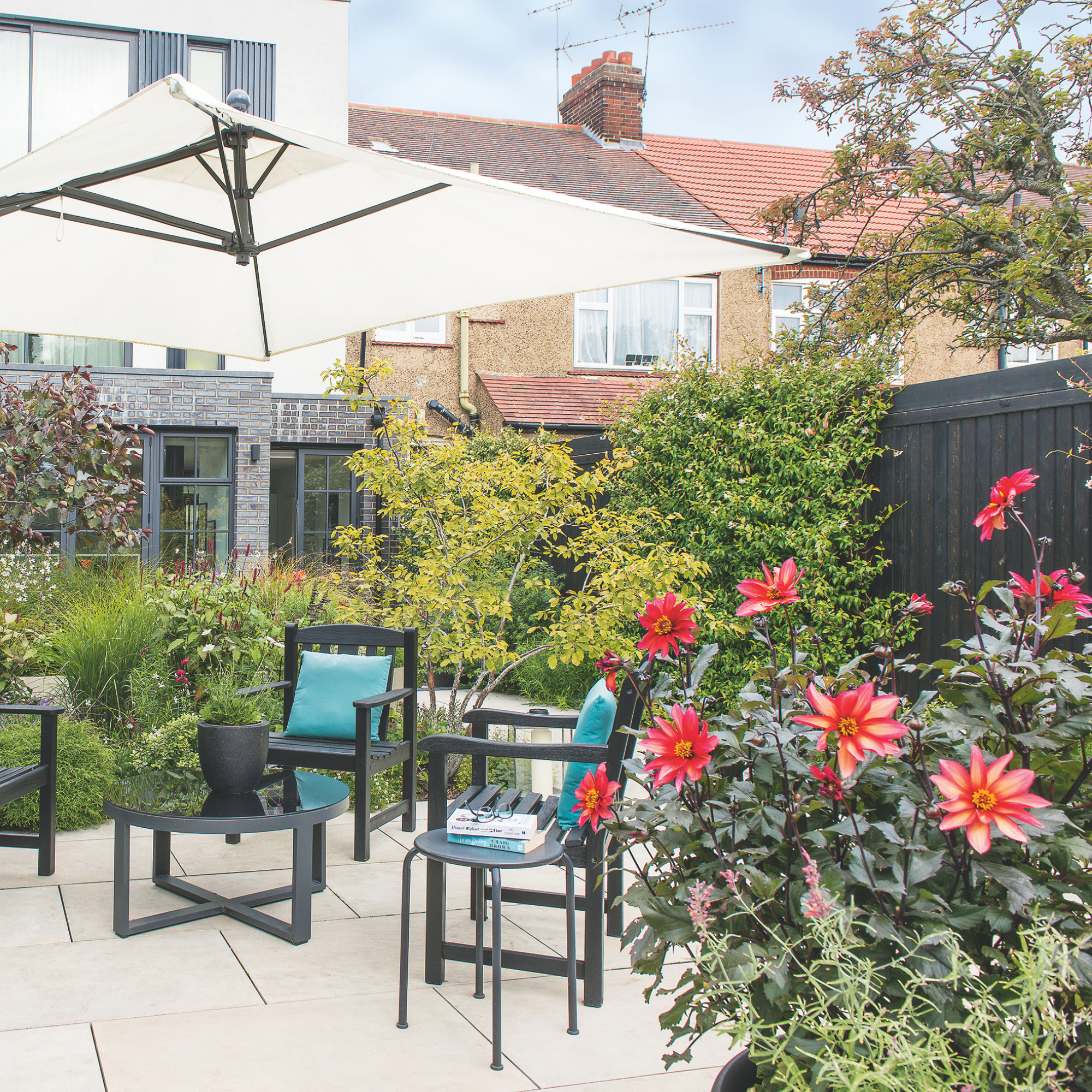 A small garden patio with tables and chairs, a sun umbrella, dense planted flower borders and flowers.