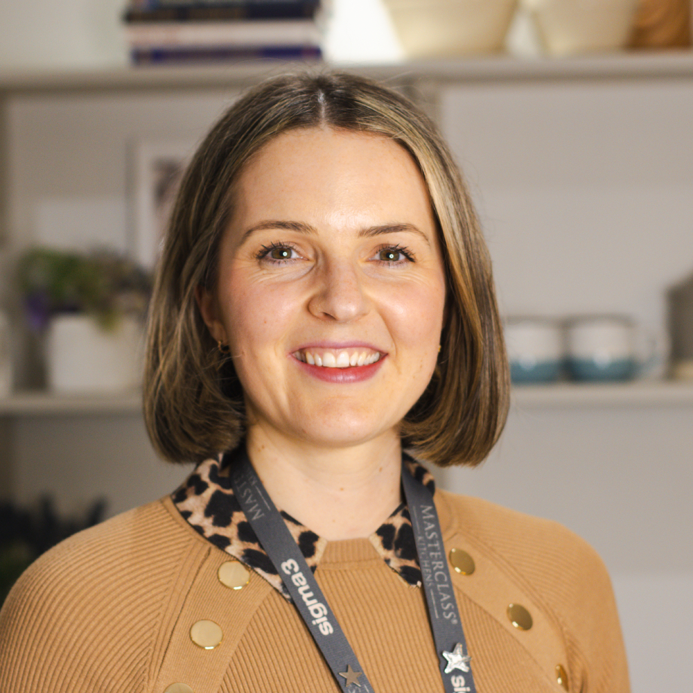 female with short light brown hair wearing tan coloured top with leopard print collar stood in kitchen