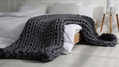 Dark gray chunky knit blanket draped over low-rise wooden bedframe with white linens on bed and wooden minimalist furniture elsewhere