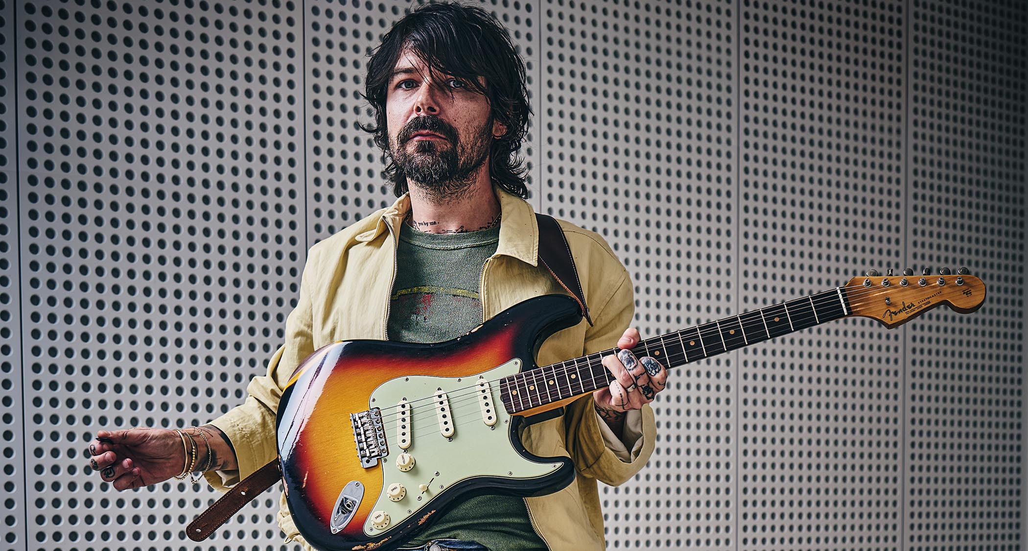 Simon Neil of Biffy Clyro wears a custard-colored jacket and green T-shirt and poses with a sunburst Fender Stratocaster