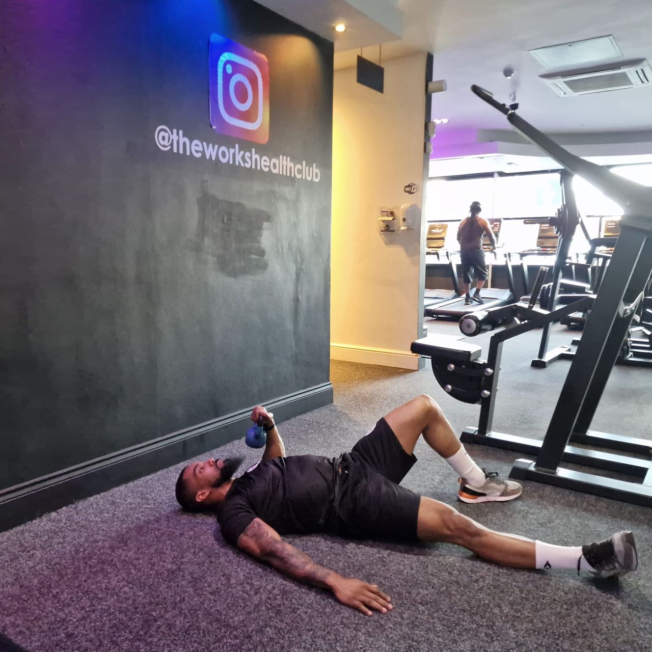 Renel Scarlett performs a kettlebell hip press inside The Works Health Club gym. He lies on the floor, with his right left straight out and left leg bent and foot planted on the floor. His right arm is on the ground, while his other arm is bent and holds a kettlebell near his shoulder. In the background we see treadmills.