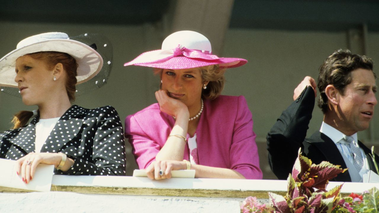 Princess Diana wearing a pink blazer and white hat resting her chin on her hand at the 1987 Epsom Derby next to Sarah Ferguson in a black polka dot coat and Prince Charles in a suit