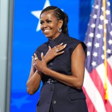 Michelle Obama wearing a sleeveless vest top. She had her hair in a braided ponytail and has her hands crossed over her chest. She is standing in front of a row of American flags. 