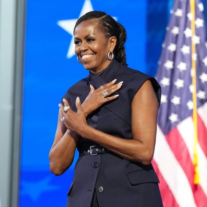 Michelle Obama wearing a sleeveless vest top. She had her hair in a braided ponytail and has her hands crossed over her chest. She is standing in front of a row of American flags.