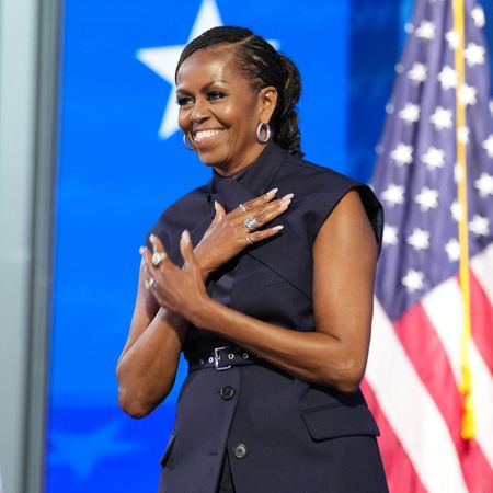 Michelle Obama wearing a sleeveless vest top. She had her hair in a braided ponytail and has her hands crossed over her chest. She is standing in front of a row of American flags. 