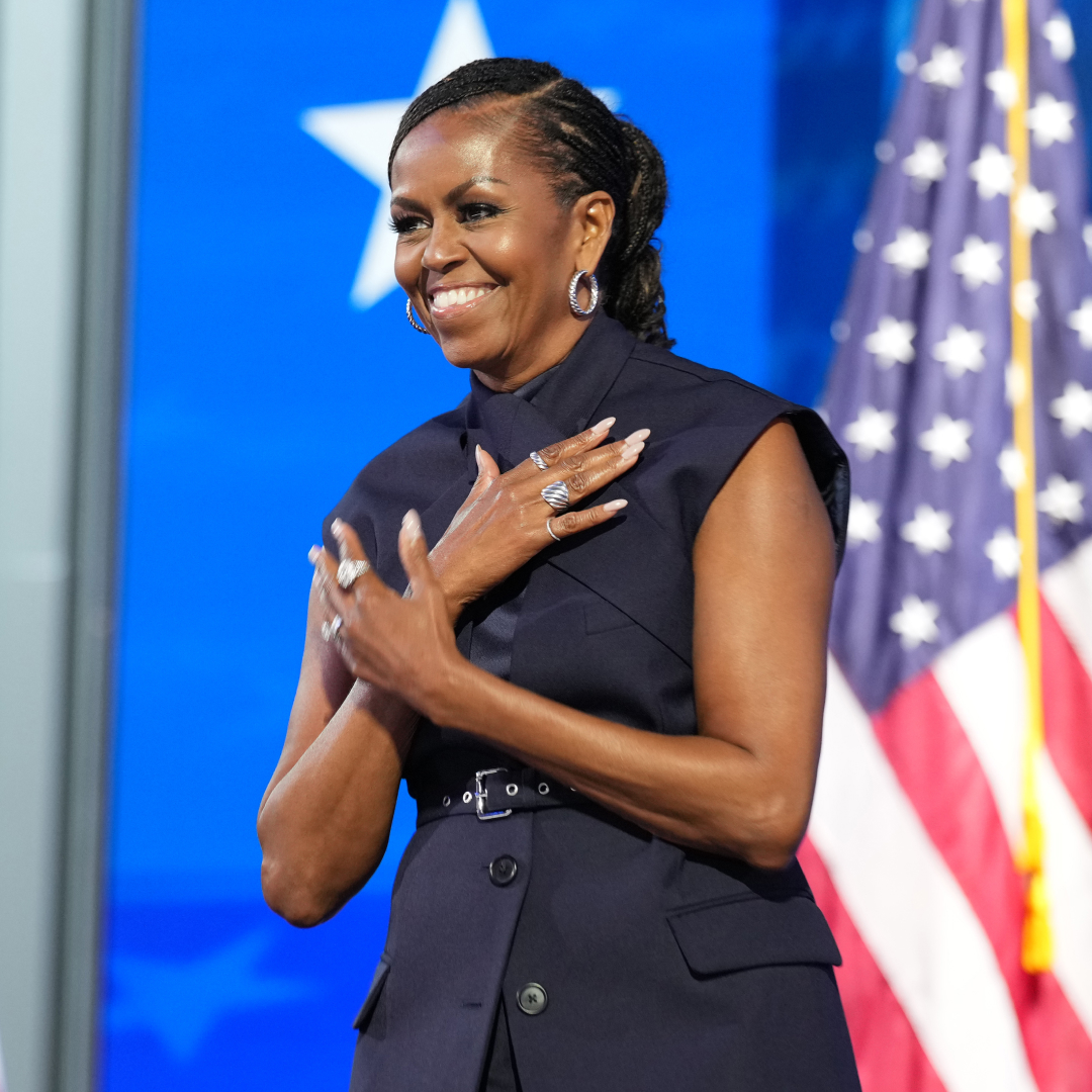 Michelle Obama wearing a sleeveless vest top. She had her hair in a braided ponytail and has her hands crossed over her chest. She is standing in front of a row of American flags. 
