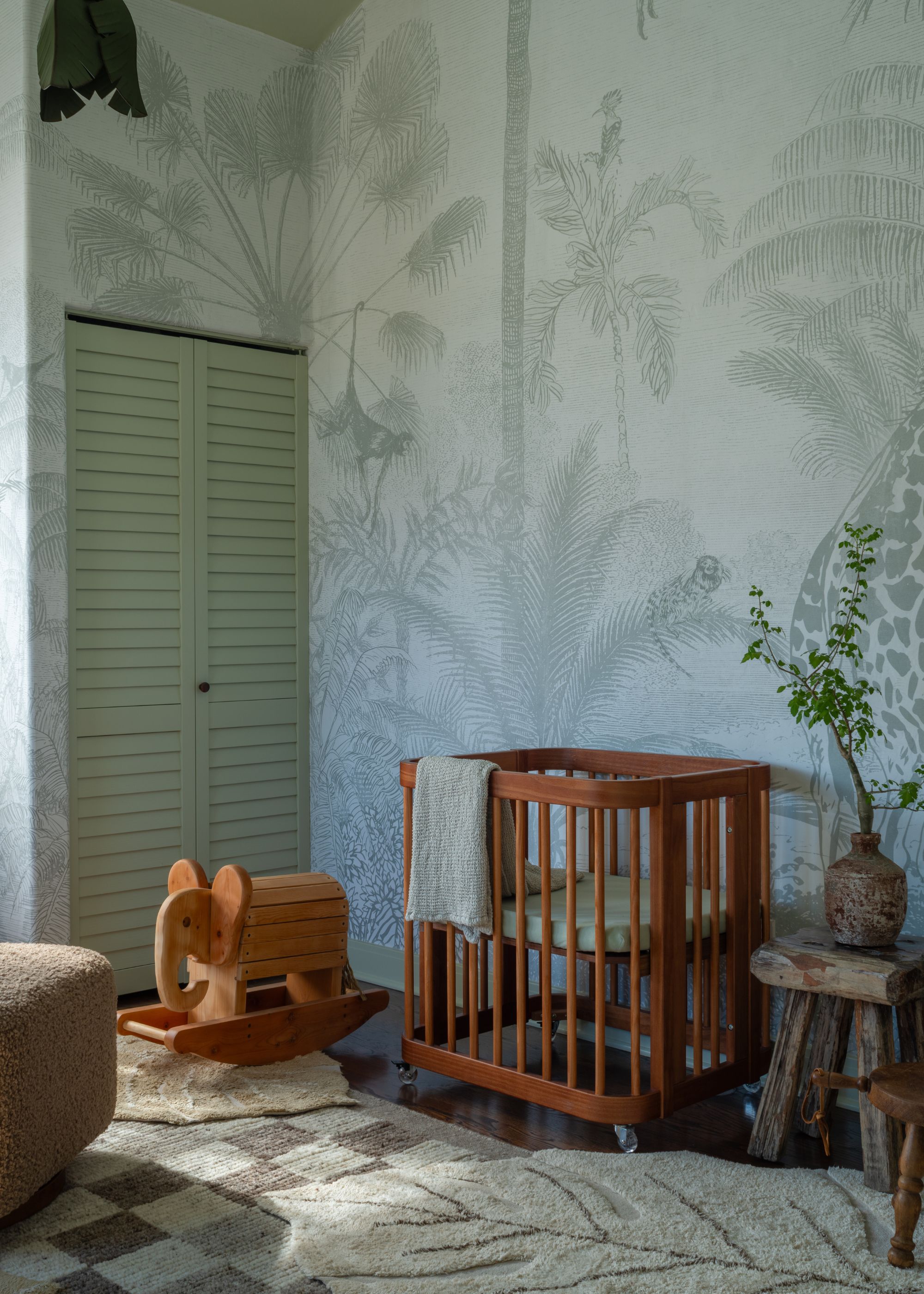 A nursery room with a botanical print wall mural in a pale green color, a pale green cupboard, a wooden cot, and neutral rugs.