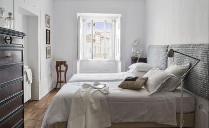 Interior view of a room at House of Birds featuring white walls, wooden flooring, two beds with pillows and white linen, two floor lamps and a dark wood chest of drawers. There is framed art on the walls and the window is open offering a view of the sky and nearby buildings