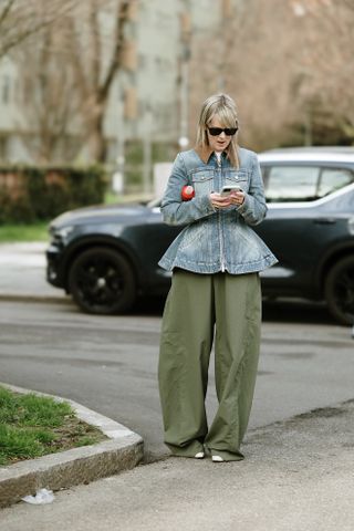 A guest at Milan Fashion Week Fall 2025 in street style wearing olive green barrel-leg pants, a blue peplum denim jacket, and black sunglasses