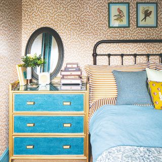 A bedroom with a patterned wallpaper and a blue chest of drawers for a bedside table next to the bed