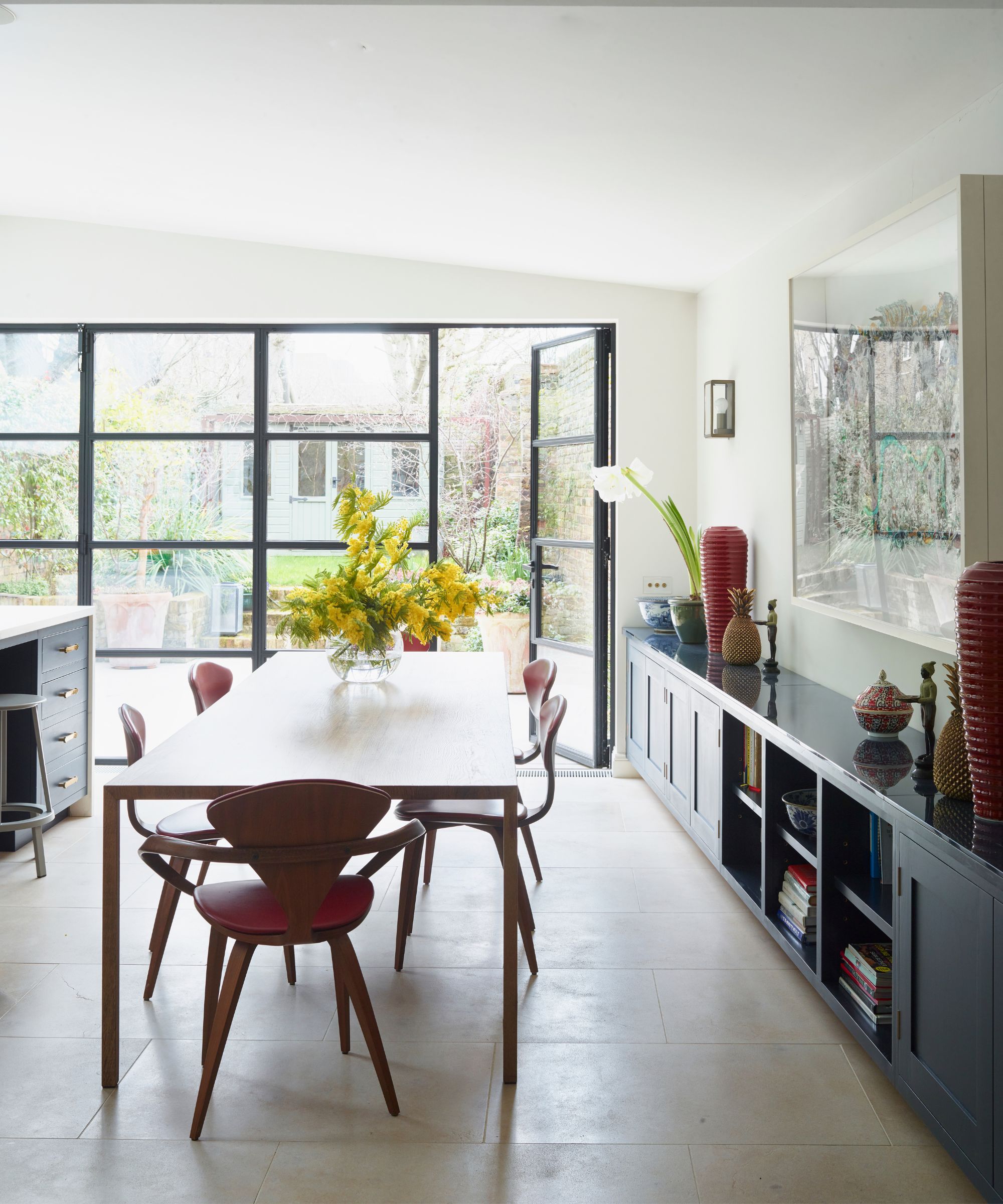 A bright kitchen dining area with tiled floors, a large wooden dining table, red chairs and blue cabinets along the right-hand side. Behind are large floor to ceiling glass doors, with one open to the right.