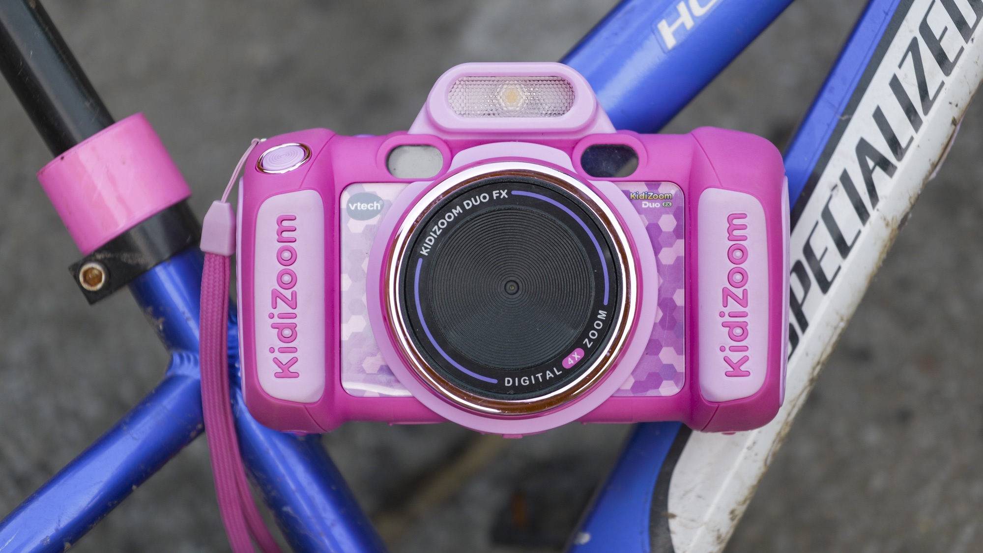 A pink V-Tech KidiZoom Duo camera resting on the blue and white frame of a kid&#039;s bike