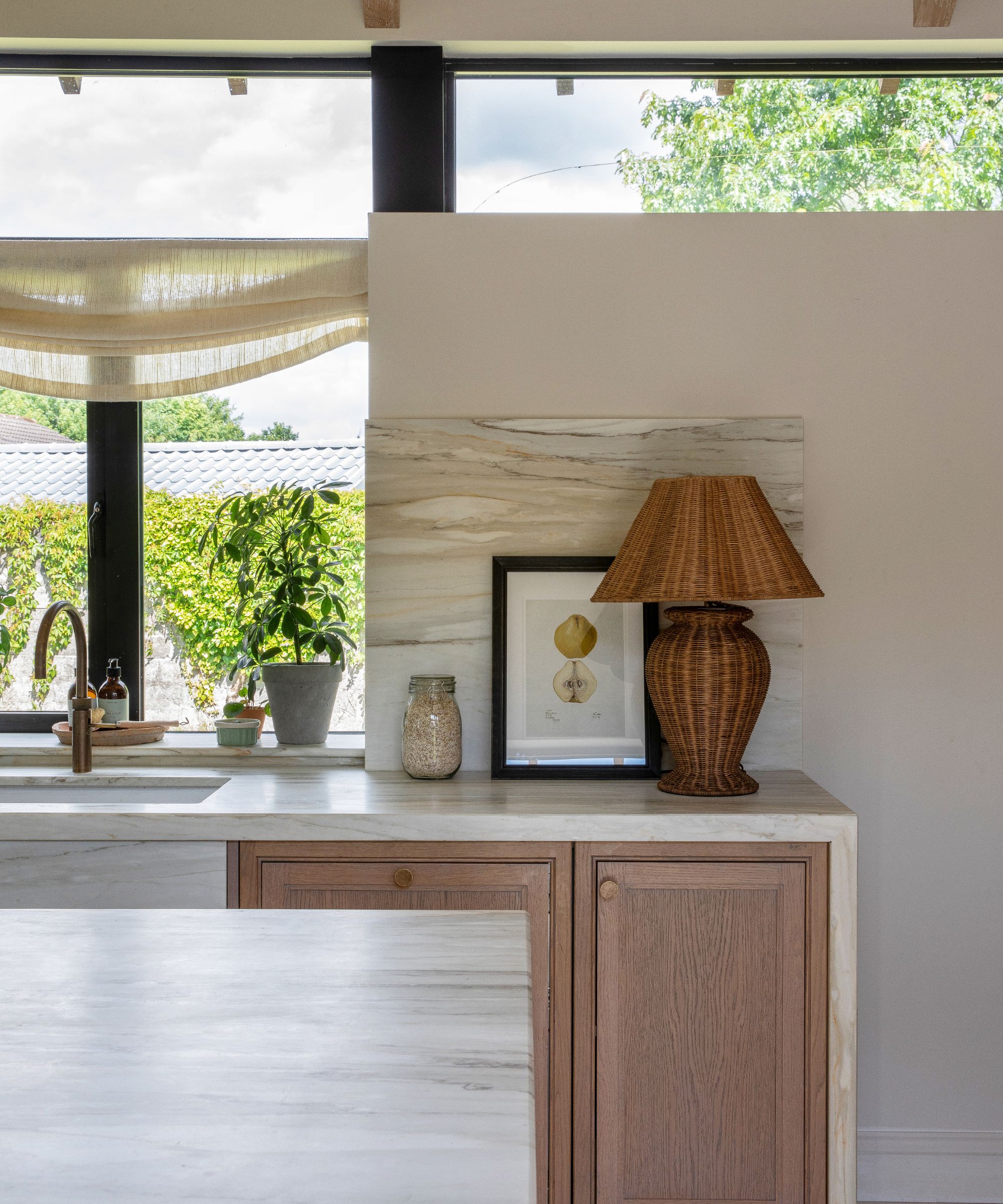 The corner of a wooden kitchen with white walls, marble countertops, and collected decor, including a rattan lamp and framed art