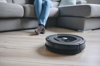 A robot vacuum in front of a sofa with the legs of a person sitting on the sofa visible