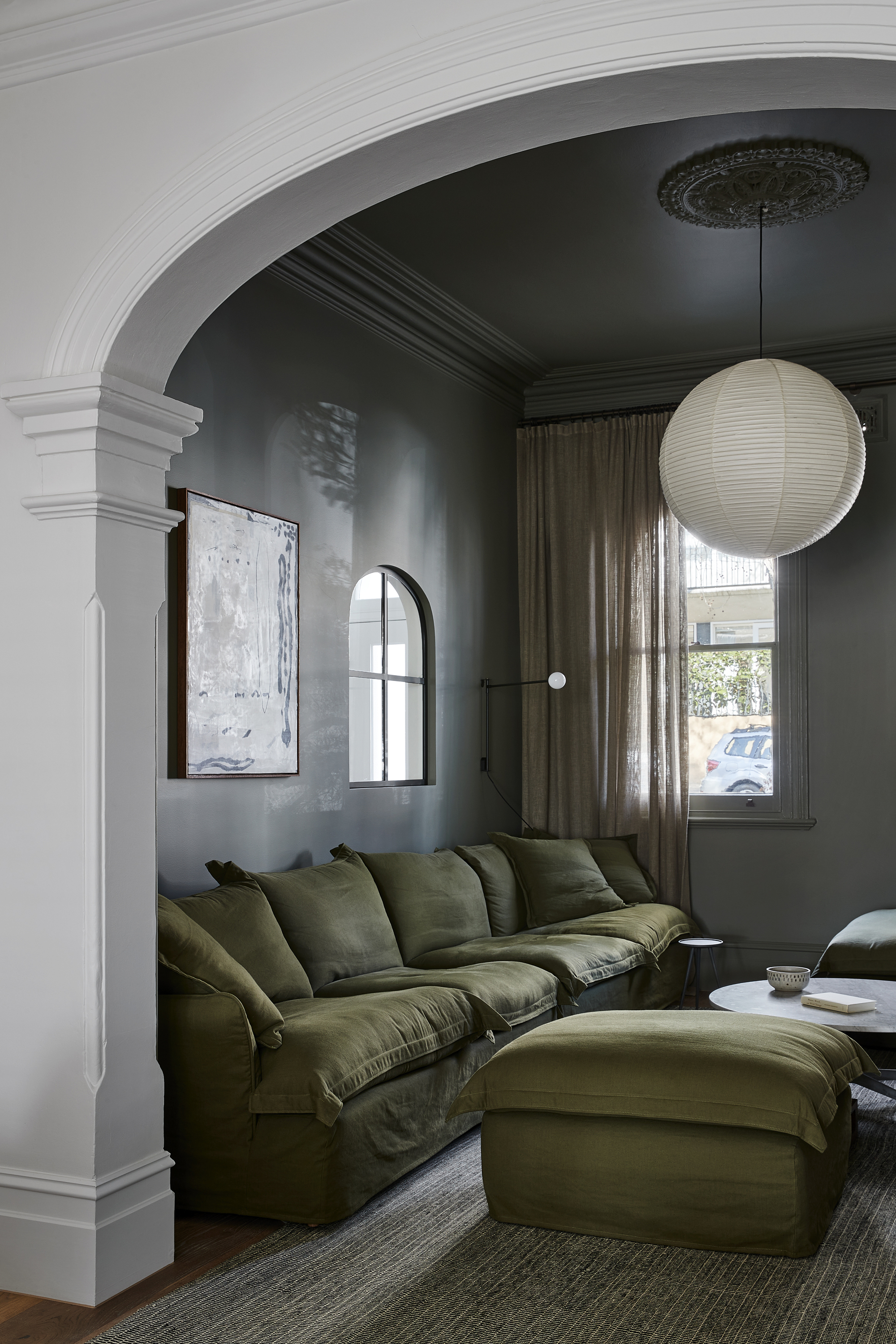 gray living room with arched feature, olive green sofa and matching ottoman, green jute rug, paper lantern pendant and windows covered with sheer linen curtains