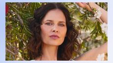 a model with clear skin and brown hair pictured outside standing in front of some foliage 
