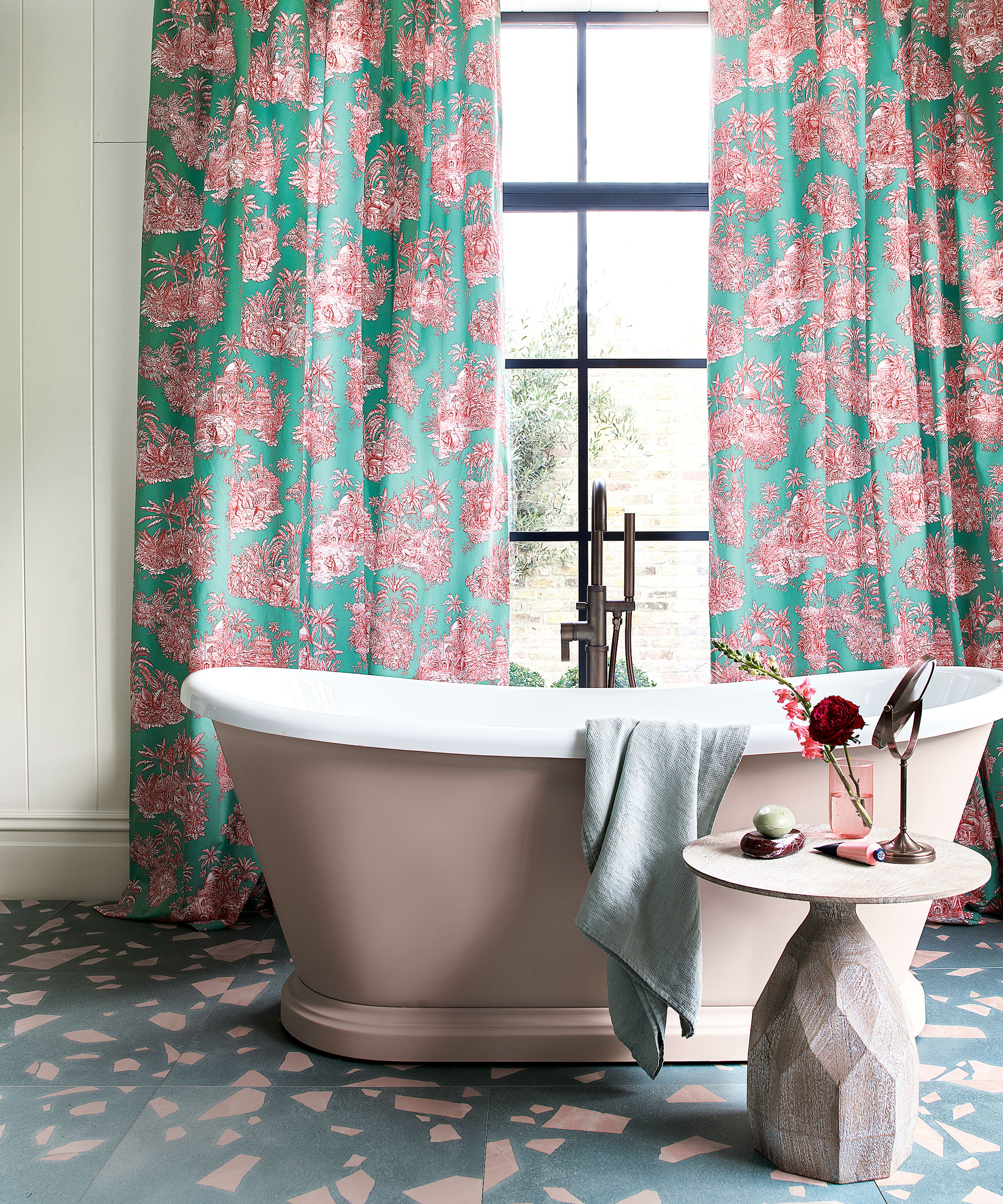 Bathroom space with pink and blue scheme, pink and blue terrazzo flooring, pink and white bathtub, pink and blue floral curtains, rounded, stone side table