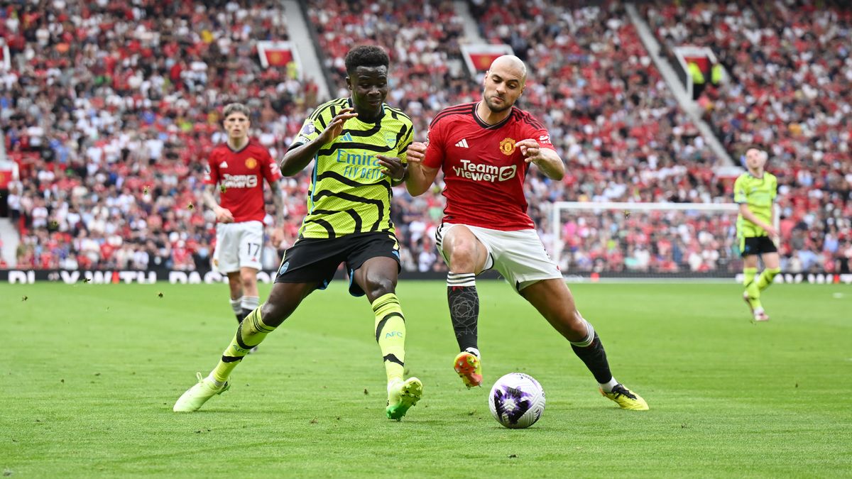 Bukayo Saka (L), wearing yellow and black strip, and Sofyan Amrabat, wearing red top and white shorts, challenge for the ball ahead of the Arsenal vs Man Utd live stream.